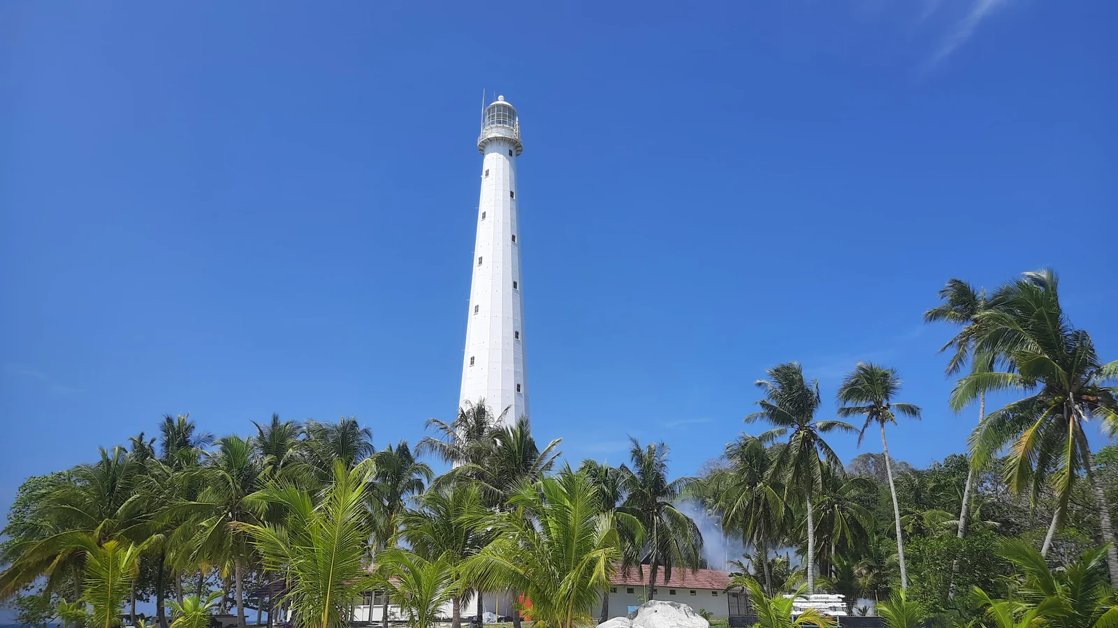 Mercusuar putih Pulau Lengkuas menjulang di atas pepohonan kelapa dan bangunan rendah di bawah langit biru.