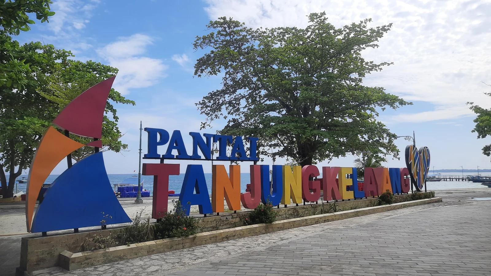 Penanda Pantai Tanjung Kelayang di sisi pelataran berpaving dan pepohonan, dengan dermaga kecil terlihat di latar kanan.